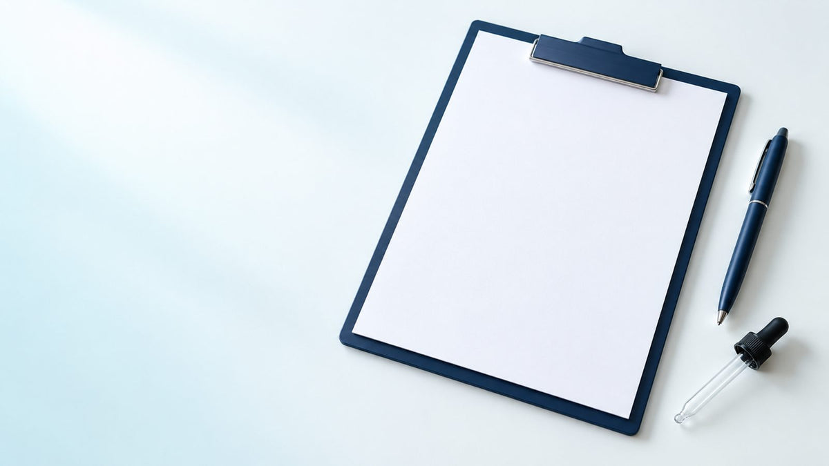 A blank sheet of paper on a navy clipboard with a navy pen and a glass dropper on a white desk