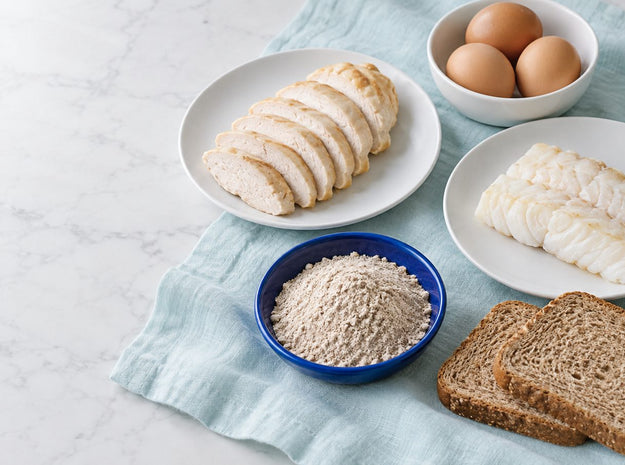 Sliced chicken, a white fish fillet, brown eggs, whole wheat bread and a bowl of flour on a pale aqua cloth