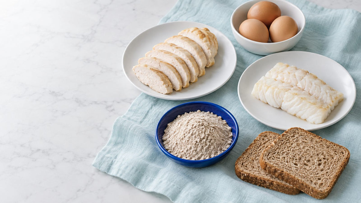 Sliced chicken, a white fish fillet, brown eggs, whole wheat bread and a bowl of flour on a pale aqua cloth